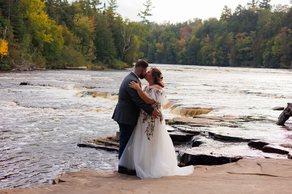 A couple in wedding attire embraces on a rocky shoreline with autumn trees and water in the background.