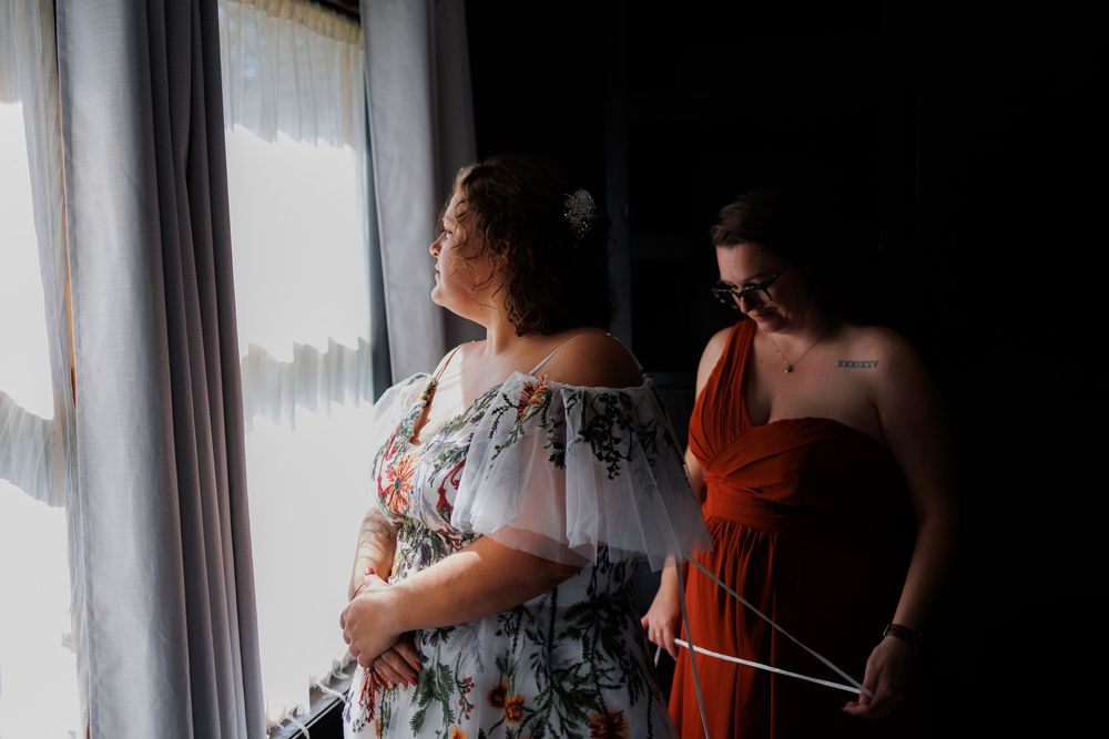 A group of people stand in a dimly lit room near windows while having a conversation.