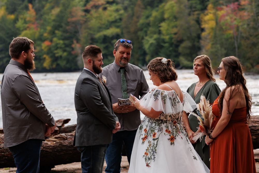 Wedding guests gather outdoors near a lake surrounded by autumn foliage during a celebration.