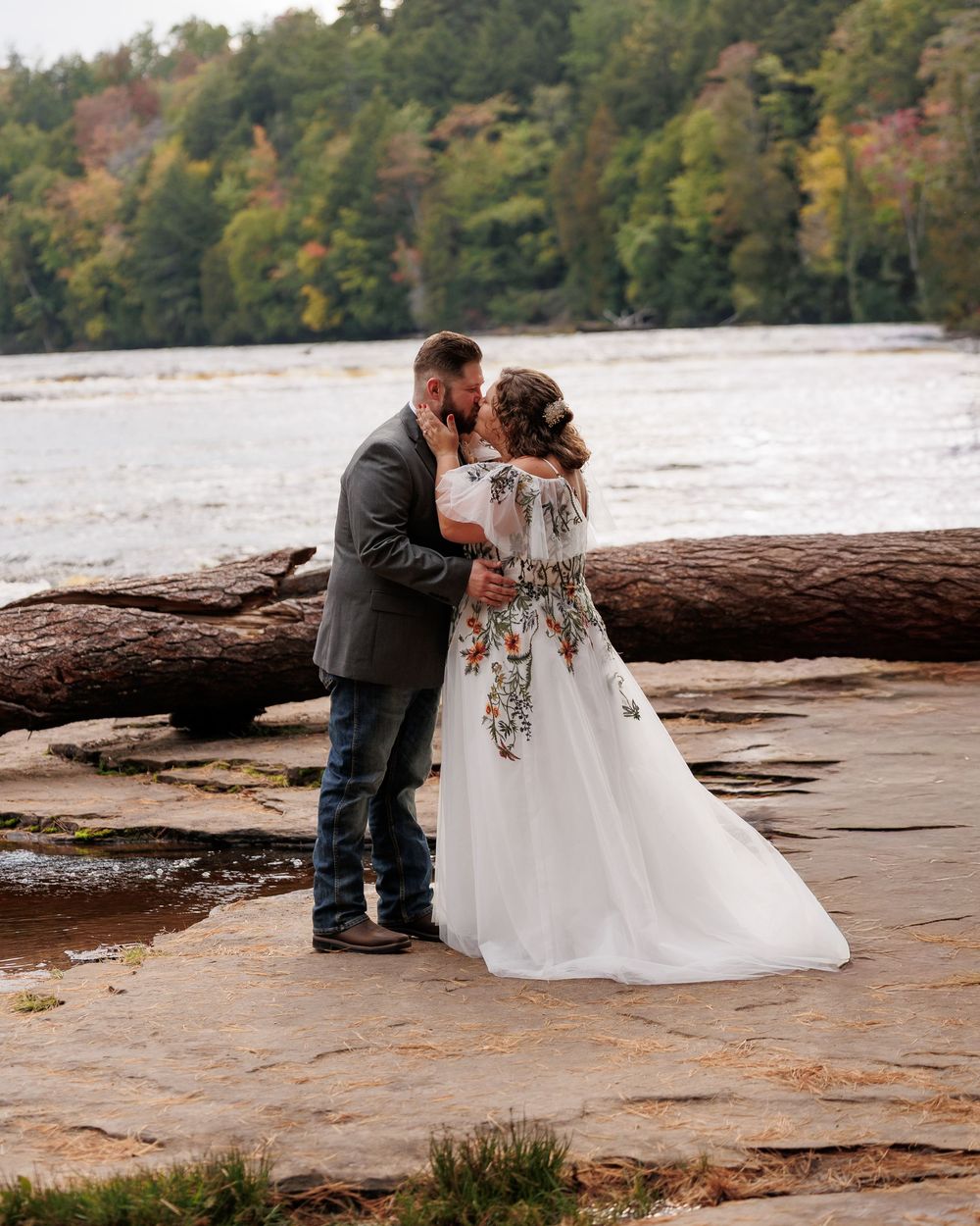 A couple shares an intimate moment on a fallen log by a lake surrounded by autumn foliage.