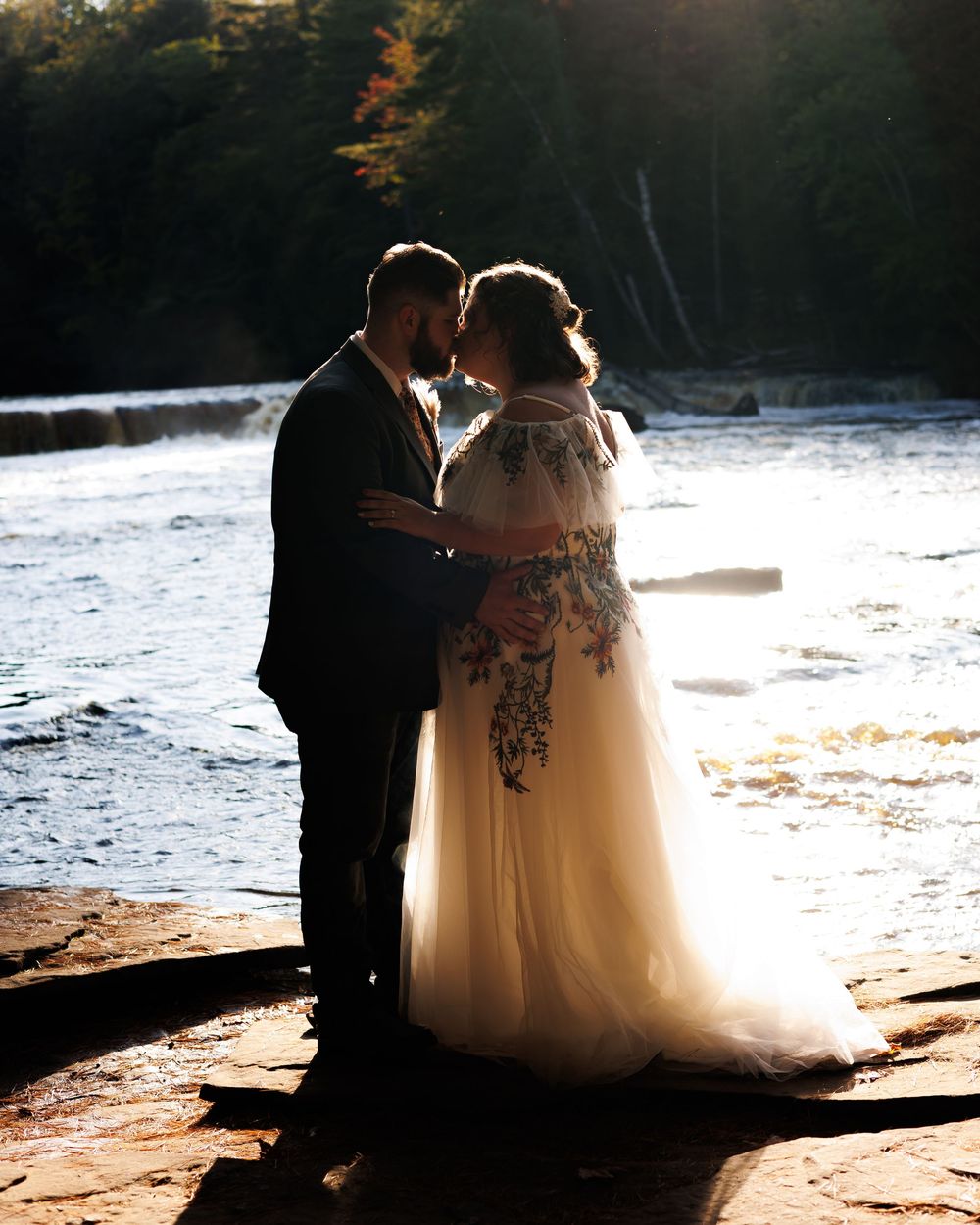 Silhouette of a bride and groom embracing by a sunlit lake during golden hour.