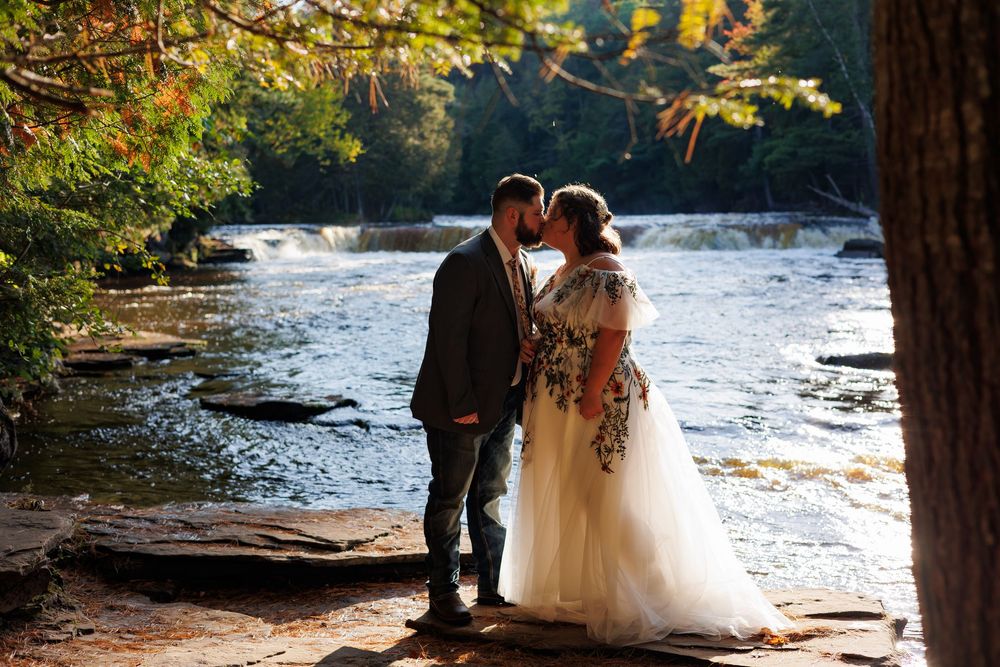 A romantic wedding couple shares a kiss by a flowing river in an autumn forest setting.