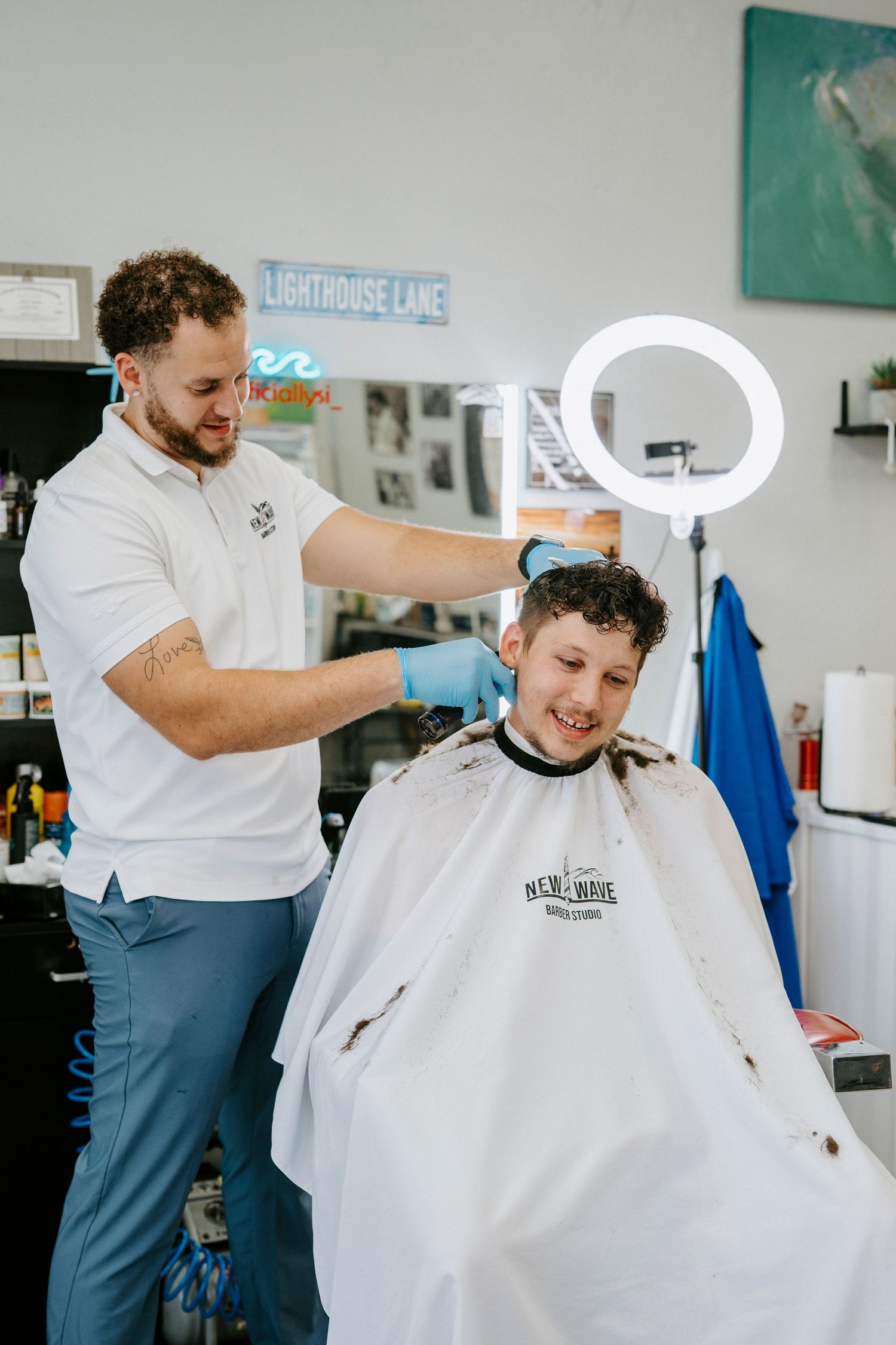A barber in a white polo shirt provides a haircut to a client in a modern barbershop setting with a ring light visible.