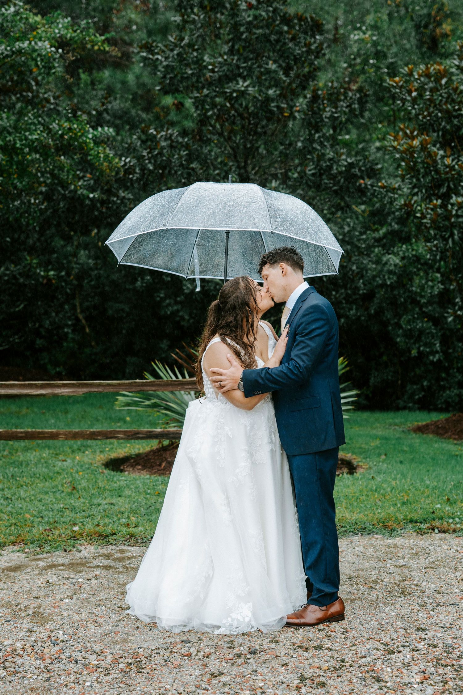 A couple shares a romantic kiss under a clear umbrella on their rainy wedding day, with lush greenery in the background.