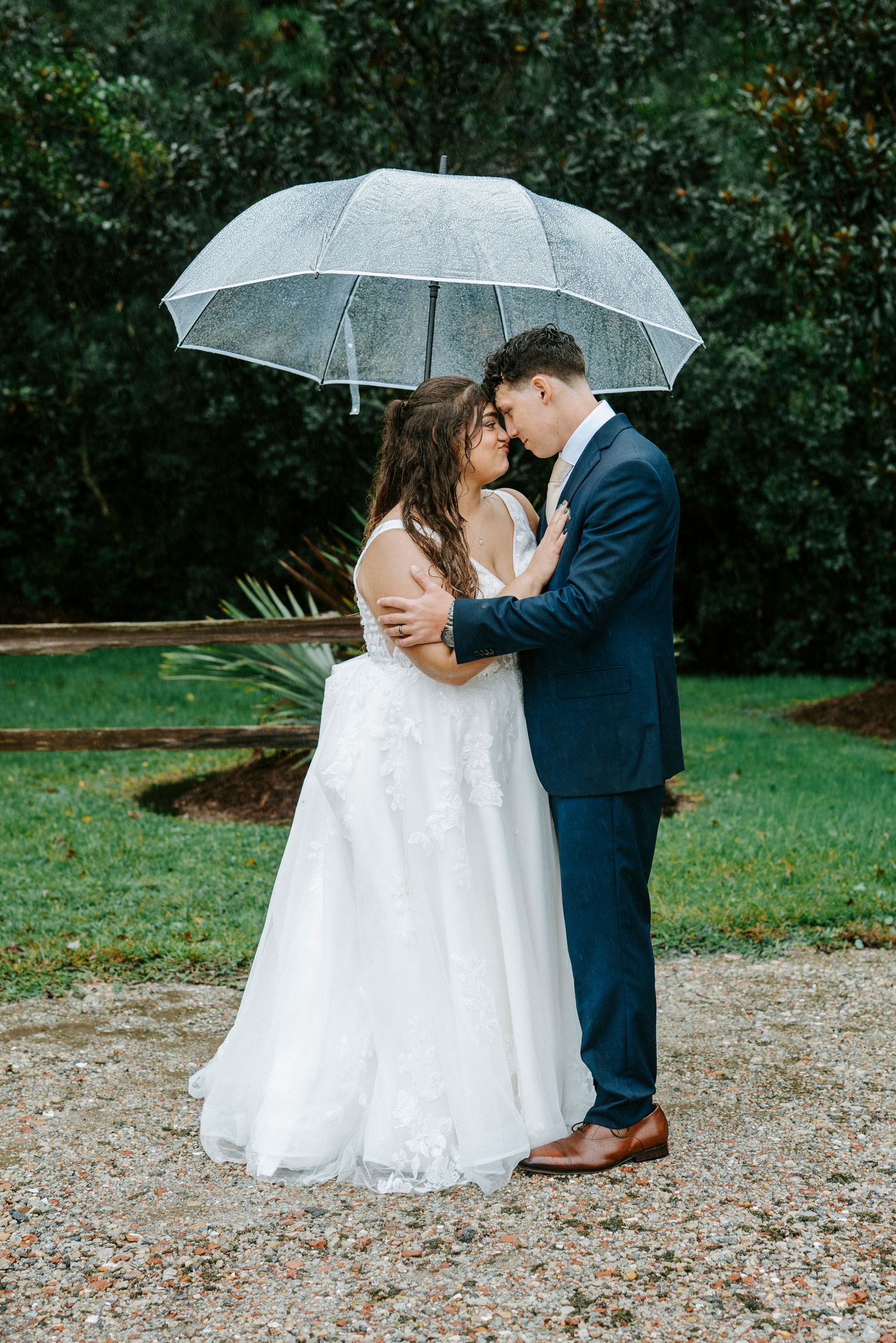 Couple shares romantic kiss under clear umbrella during outdoor wedding photoshoot in garden setting.