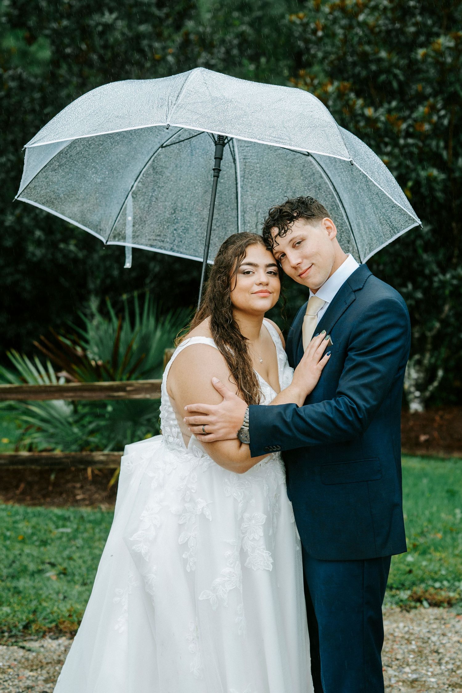 A romantic wedding couple shares an intimate moment under a clear umbrella in a garden setting.