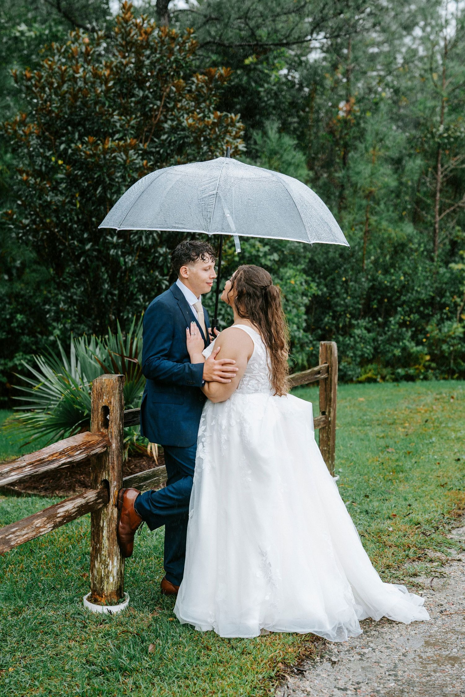 A romantic wedding couple shares intimate moments on a rustic wooden bench in the rain under a clear umbrella.