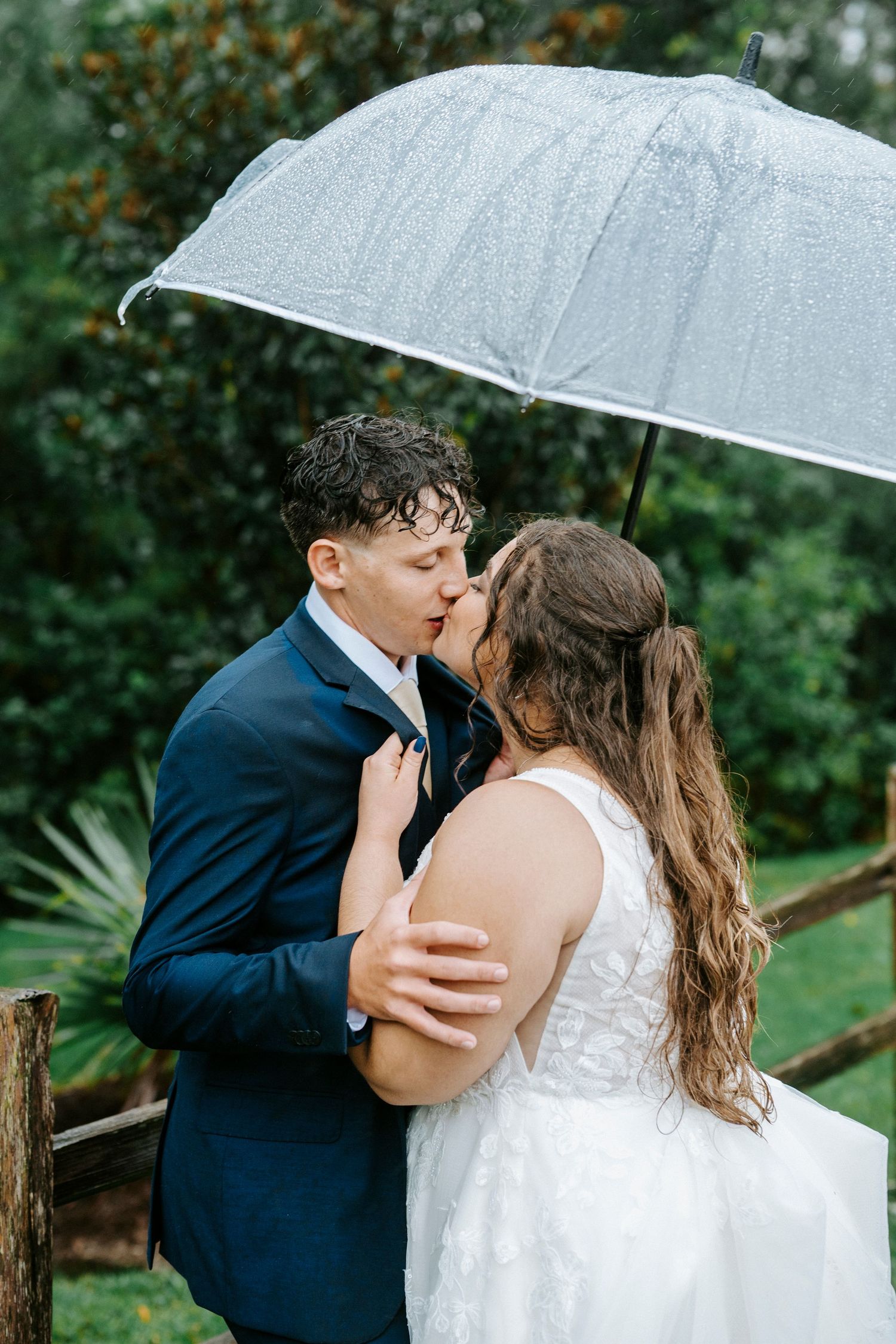 A couple shares a romantic kiss under a white umbrella in a garden setting on their wedding day.