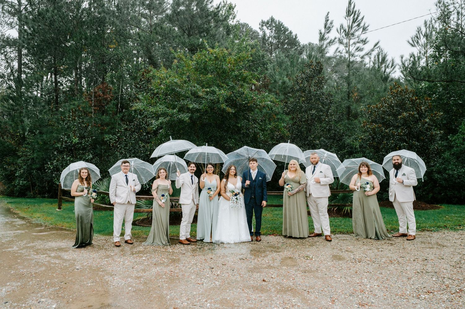 Wedding party holding white umbrellas in a line pose together on a rustic outdoor path surrounded by pine trees.