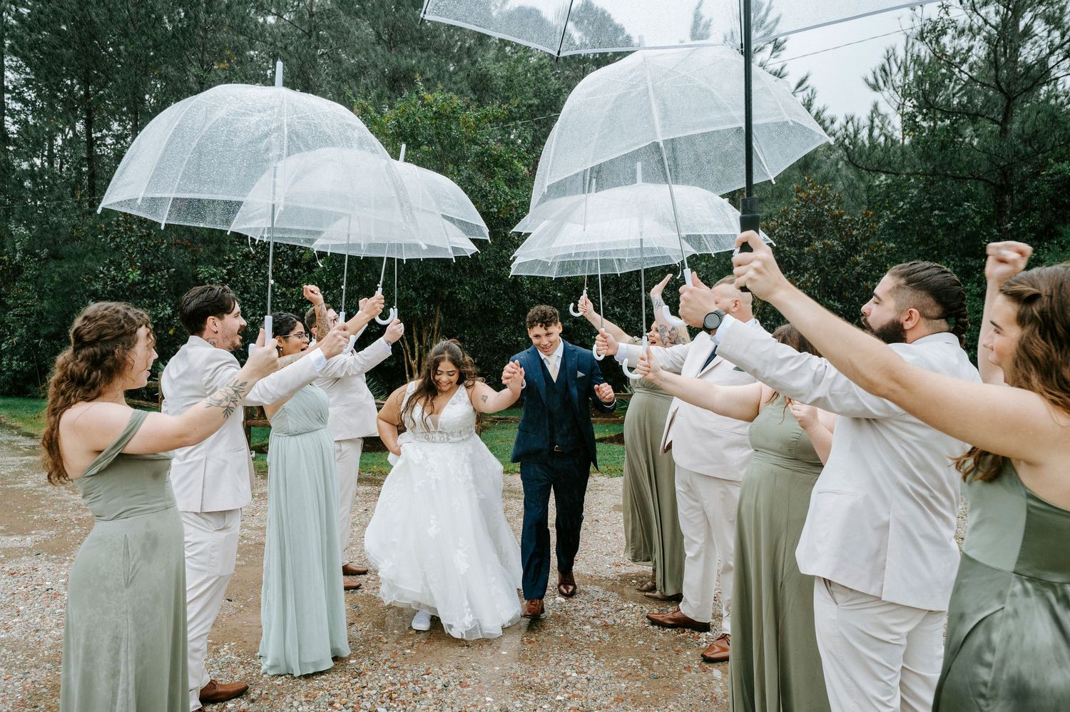 Wedding party members hold clear umbrellas while posing together in light colored dresses on a rainy day outdoors.