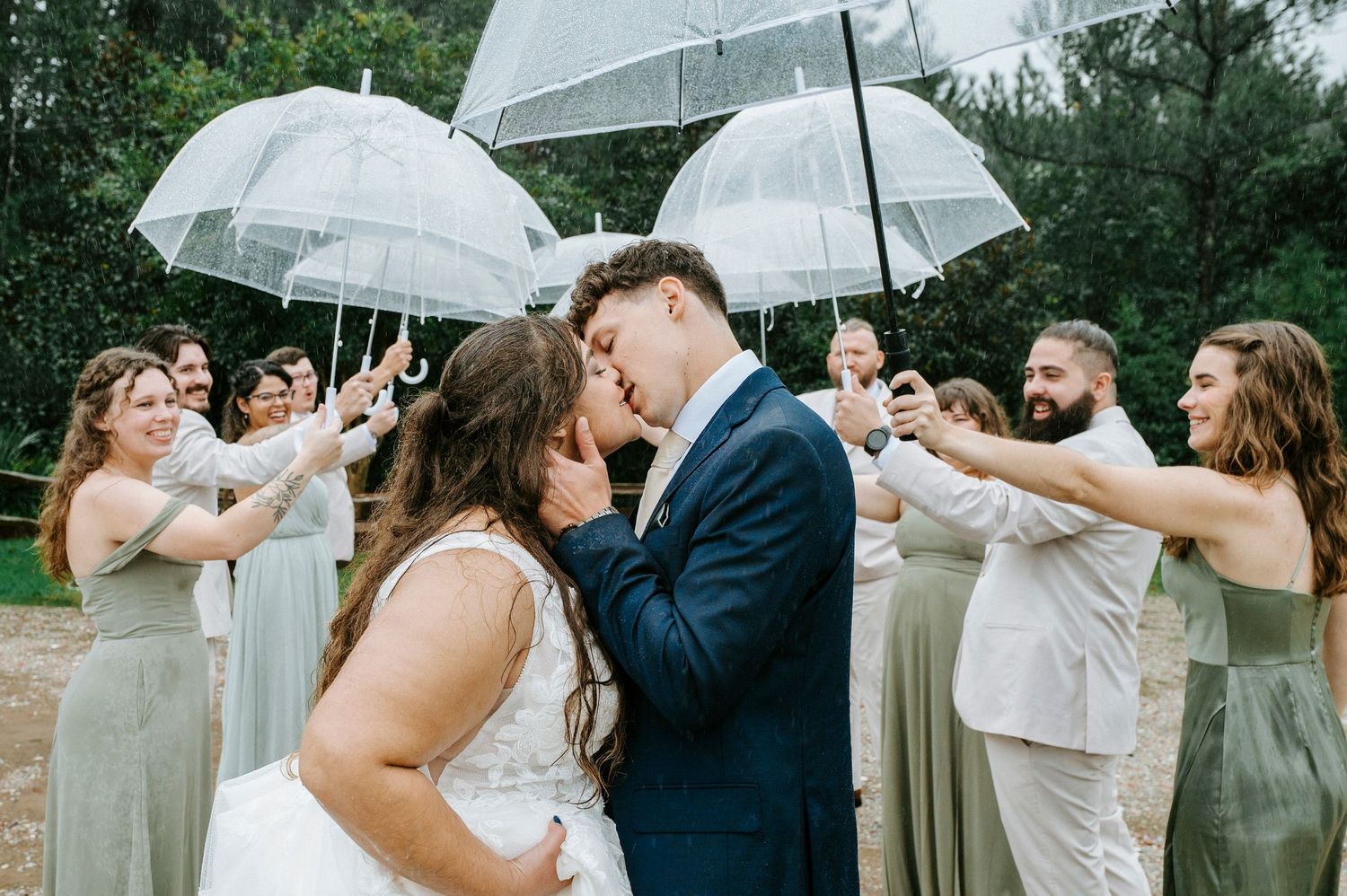Wedding party members dance and celebrate with white umbrellas in the rain during an outdoor reception.