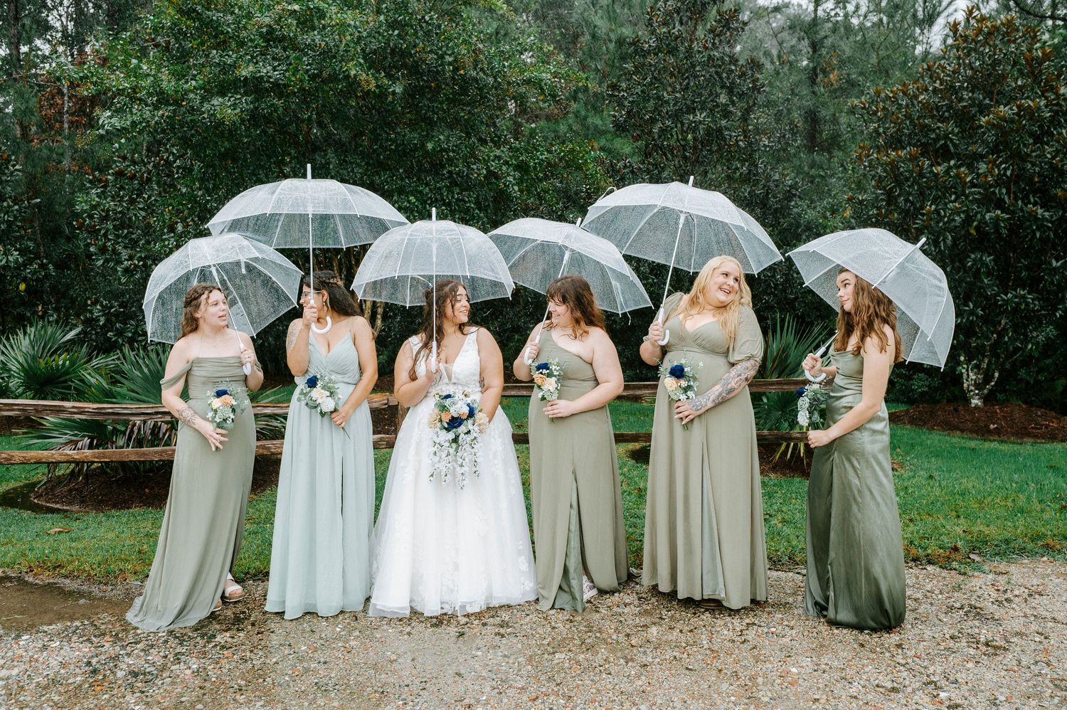 Wedding party stands in line holding clear umbrellas on a rainy day, creating an elegant outdoor photo composition.