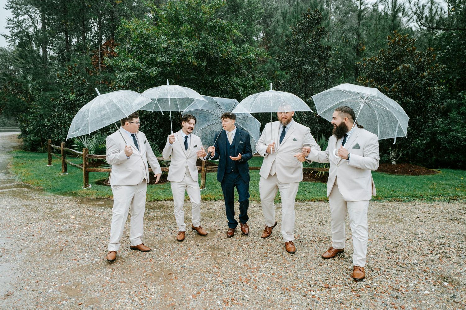 Wedding party members in white attire walk together holding umbrellas on a rainy day at an outdoor venue.