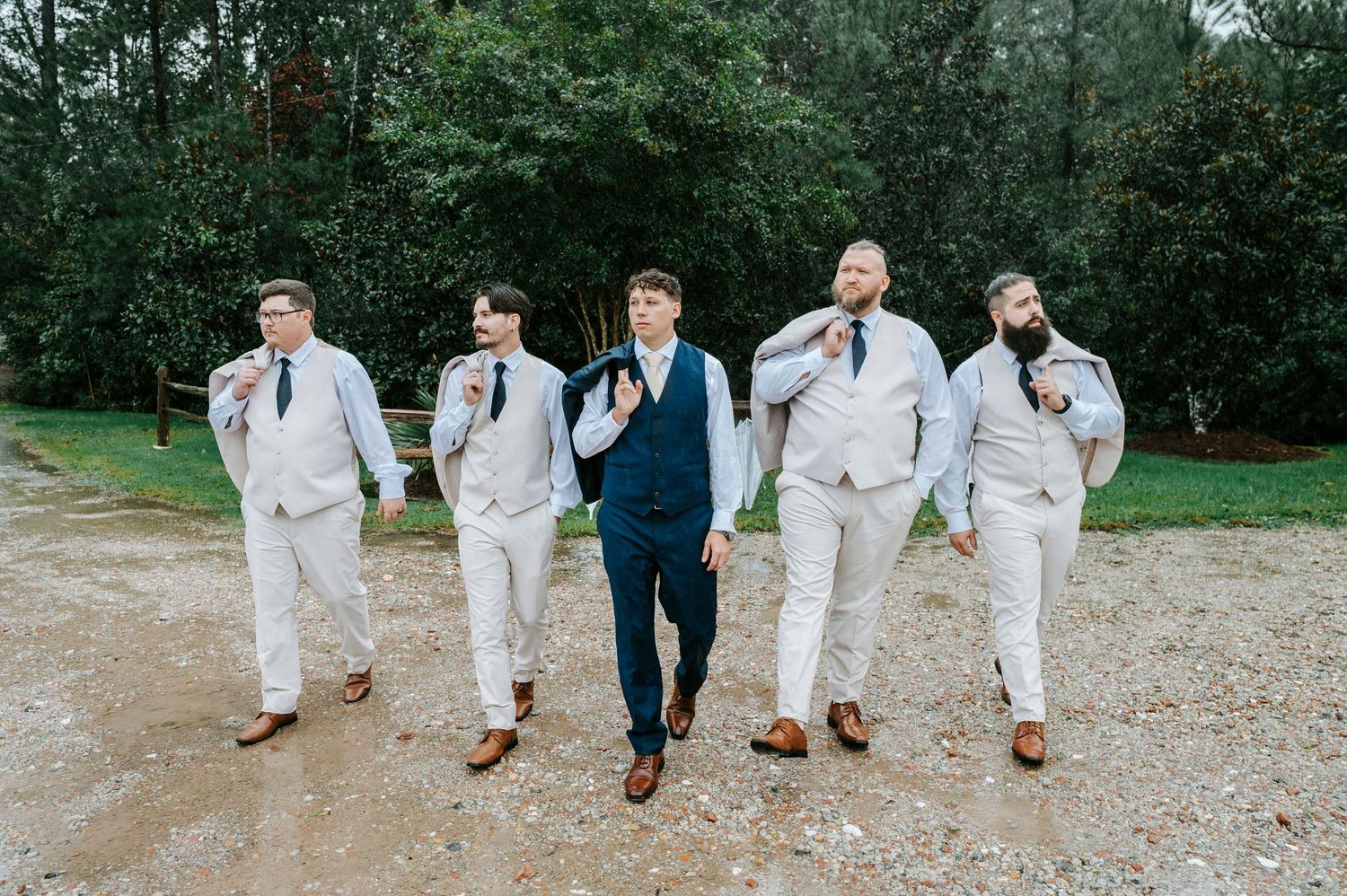 Groomsmen in white suits and navy blue jackets walk together on a gravel path during a wedding celebration.
