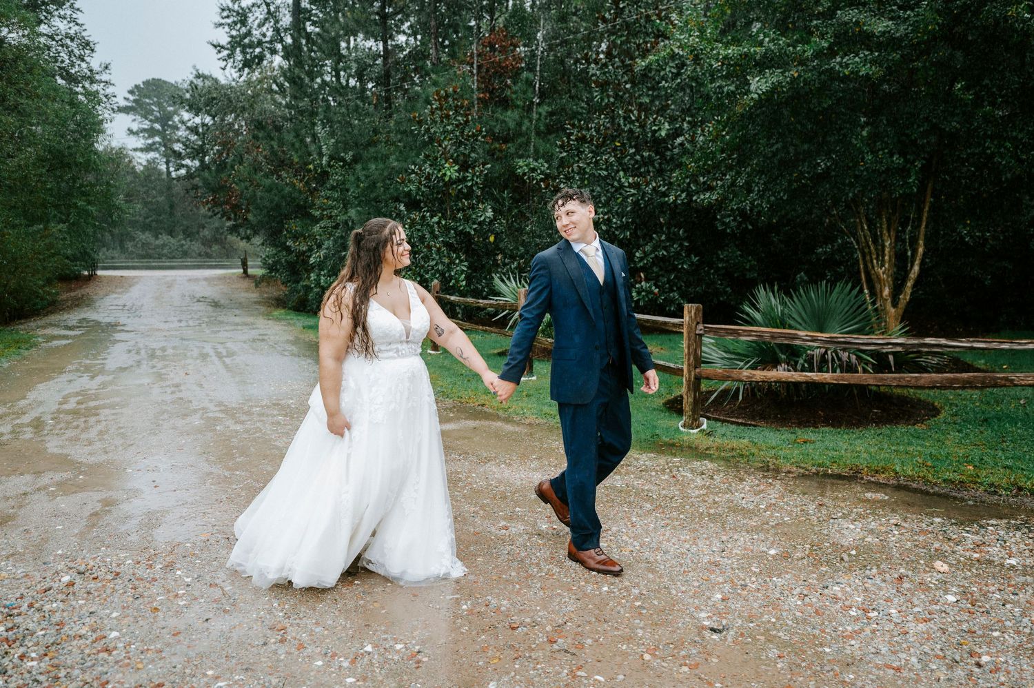 A couple walks along a gravel path surrounded by lush green trees during a wedding photoshoot.
