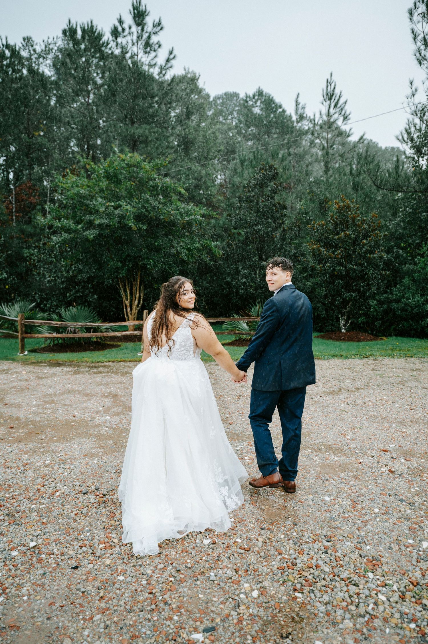 A couple in wedding attire dances together on a gravel path surrounded by pine trees.