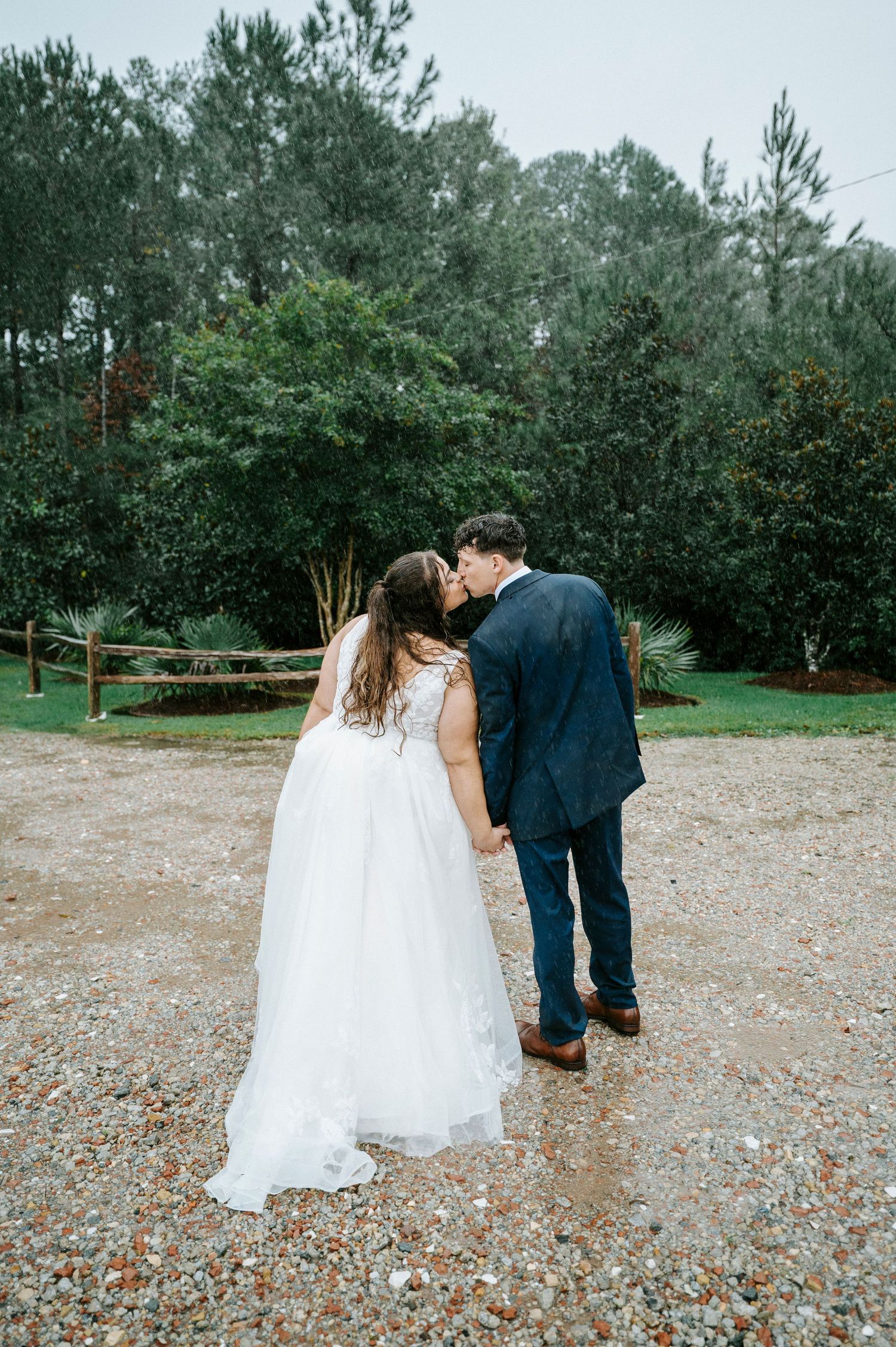 A romantic wedding moment captured from behind as the couple stands on a gravel path near pine trees.