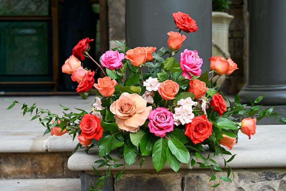 A vibrant floral arrangement of peach, pink and coral roses with green foliage displayed on a stone ledge.