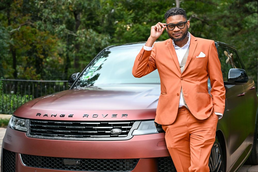 A person in a bright orange suit poses with a Range Rover vehicle in an outdoor setting.