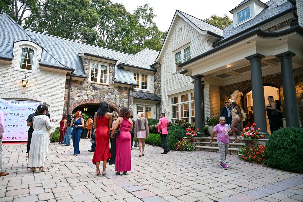 Guests in colorful formal attire gather outside a stone and white mansion during an elegant outdoor event.