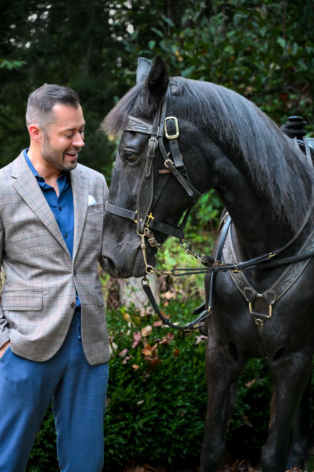 A person in a gray vest and blue shirt interacts affectionately with a black horse in a garden setting.
