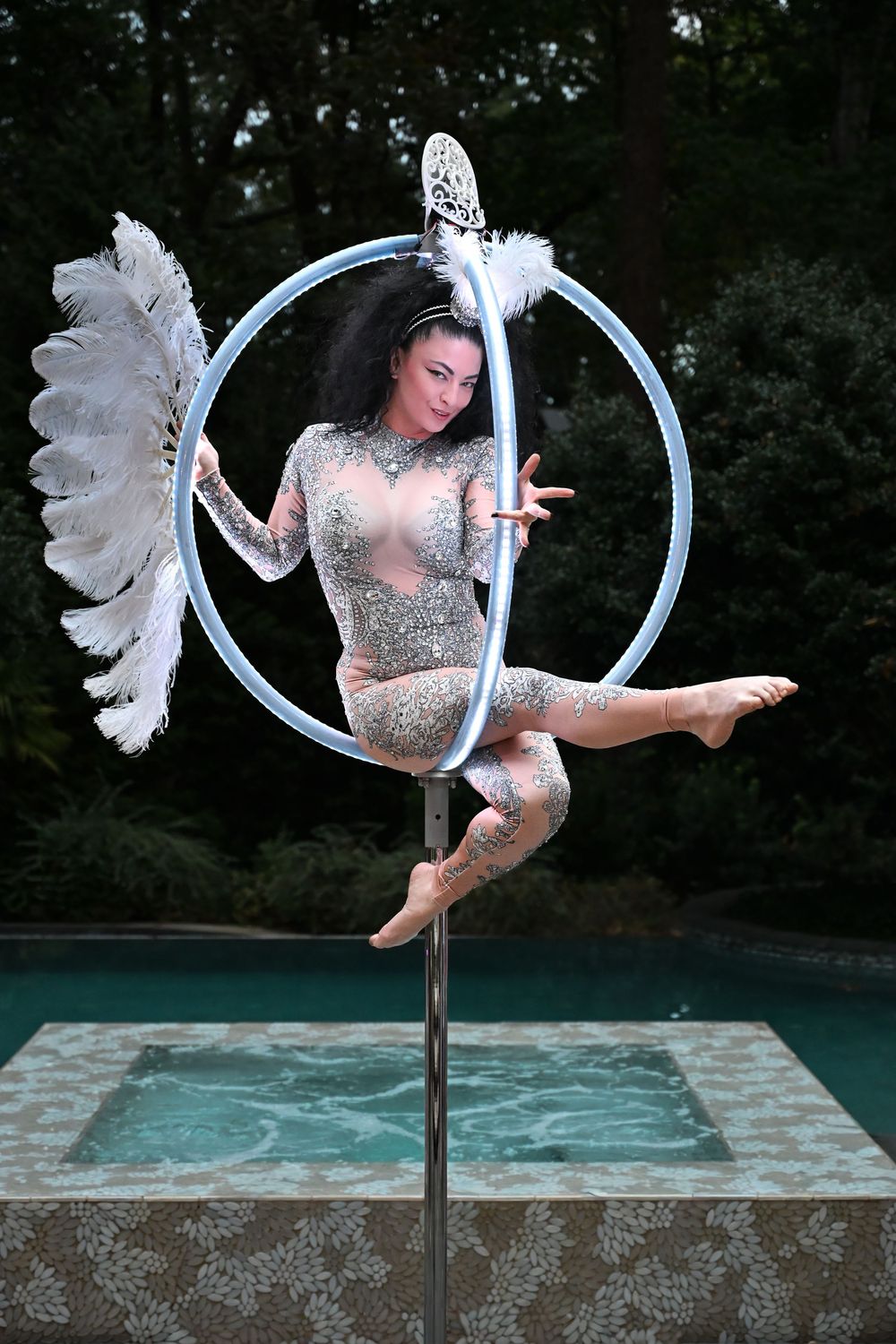Aerial performer in sparkly costume gracefully poses on lyra hoop with white wings above a pool.
