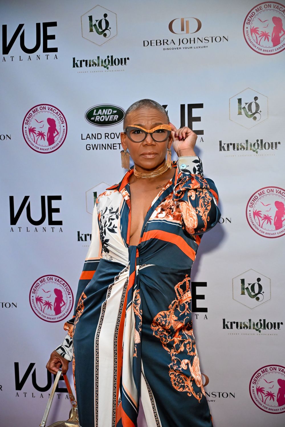 A person poses at a red carpet event wearing a black and orange patterned outfit against a backdrop with sponsor logos.