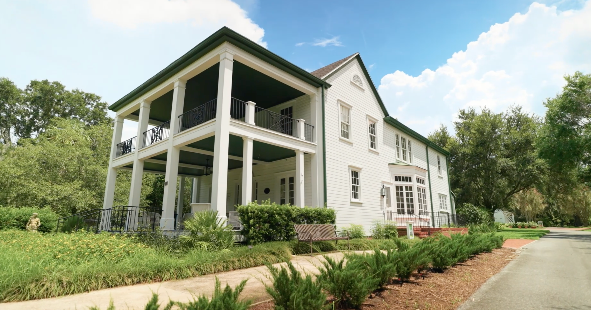 A modern white farmhouse style home with a wraparound porch sits nestled among lush green landscaping.
