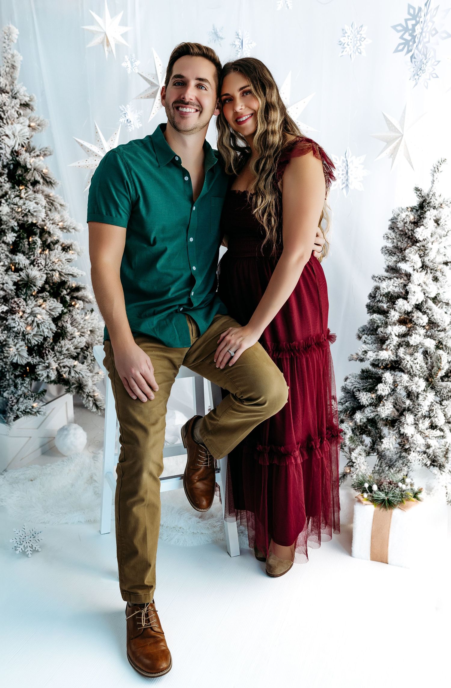 A couple poses in holiday attire against a winter wonderland backdrop with white trees and snowflake decorations.