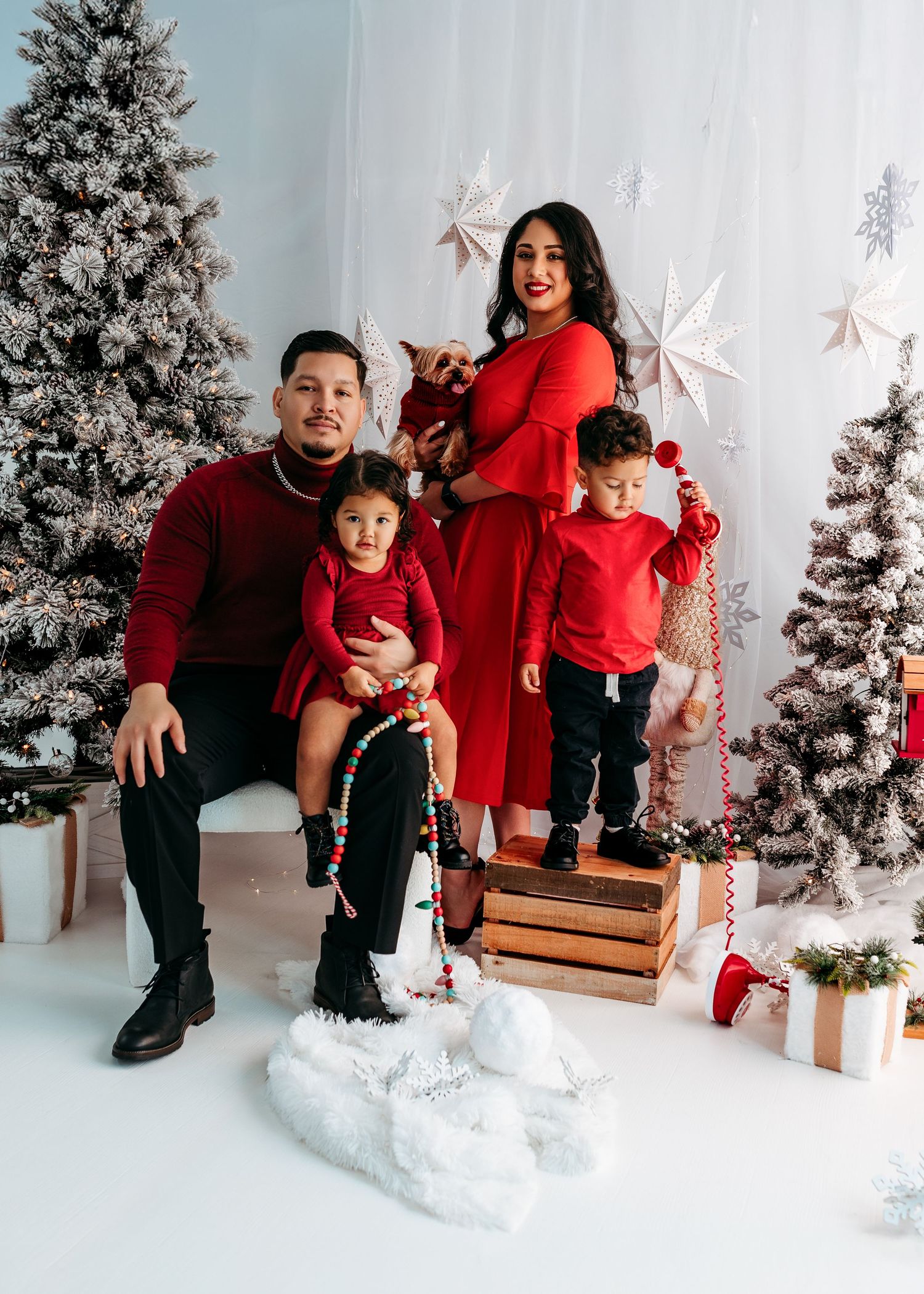 A family wearing coordinated red and black outfits poses in a winter wonderland setting with snow and Christmas trees.