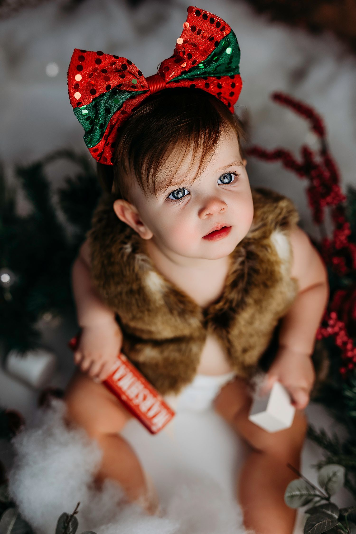 A small child wearing a red and green holiday bow sits among white cotton snow and festive red berries for a Christmas photo.