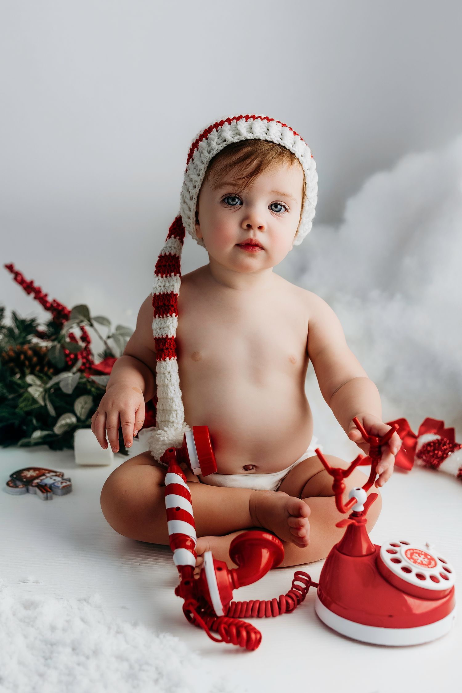 A baby wearing a red and white striped winter hat sits on white bedding with a vintage red toy telephone nearby.