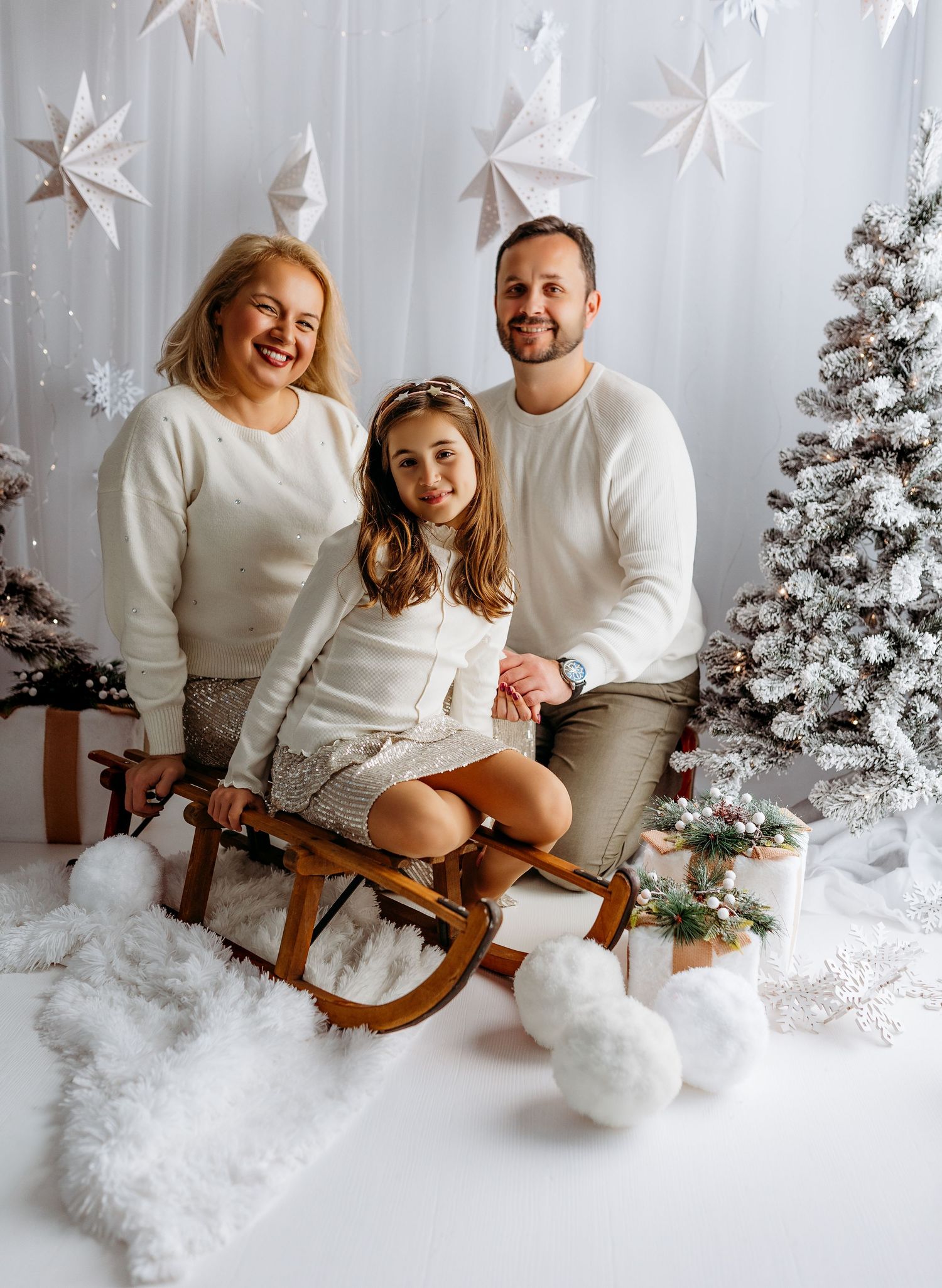 A couple in white sweaters sits on a vintage wooden sleigh against a snowy winter backdrop with Christmas decorations.