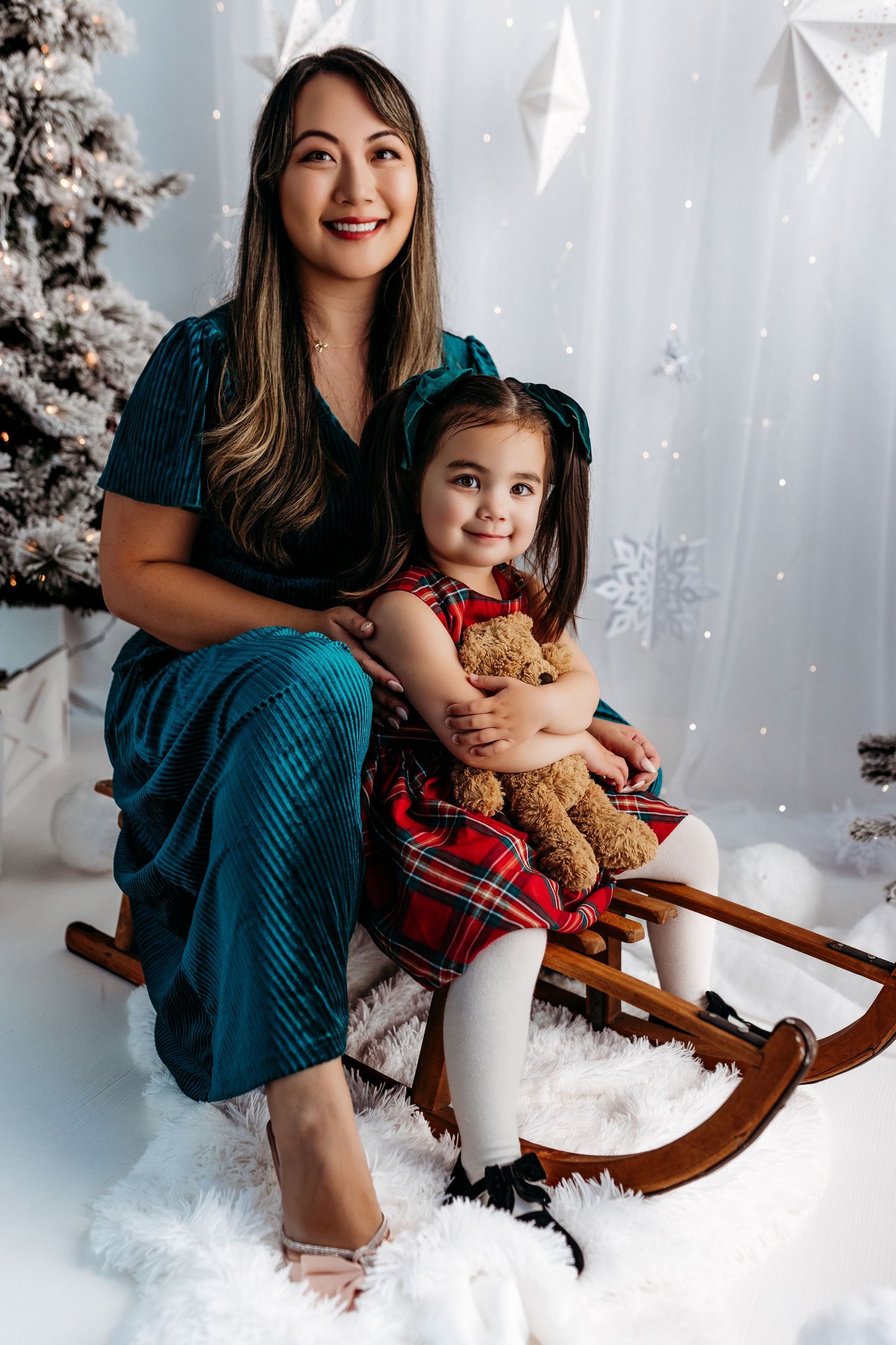 Mother and daughter in winter holiday setting pose together on wooden sleigh with teddy bear and plaid blanket.