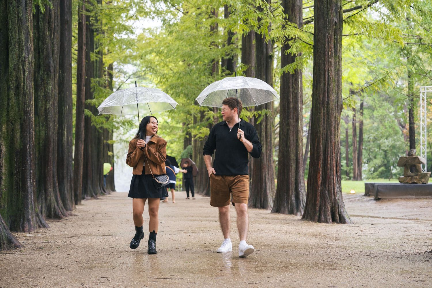 People with umbrellas walking along a tree-lined path in a park on a rainy day.