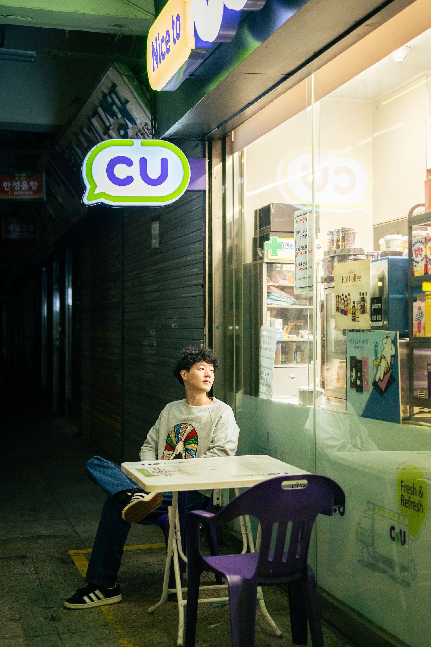 A person sits at a table outside a CU convenience store at night with bright storefront lighting.