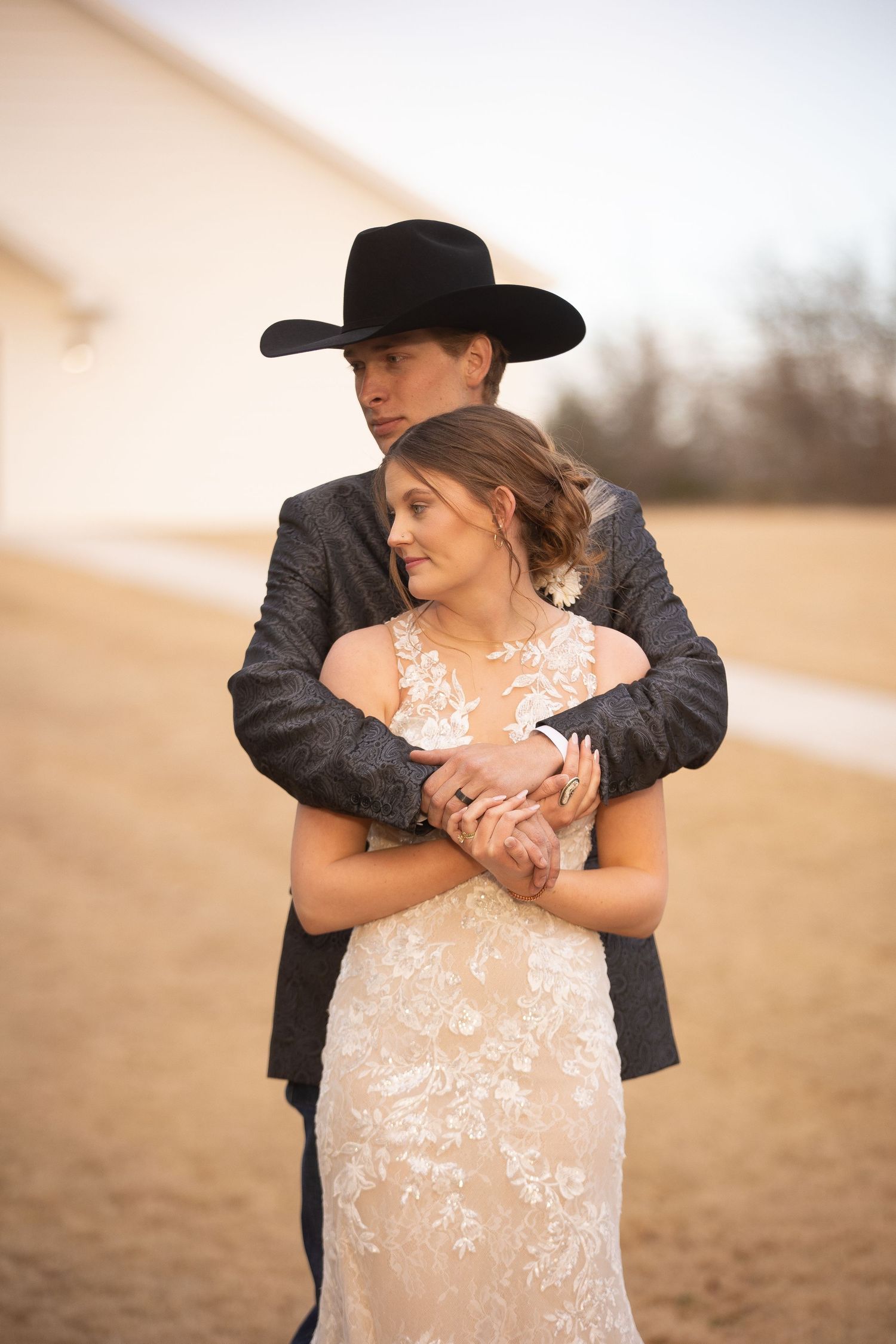Romantic couple embraces on a sandy beach at sunset with person wearing black cowboy hat and white lace dress.