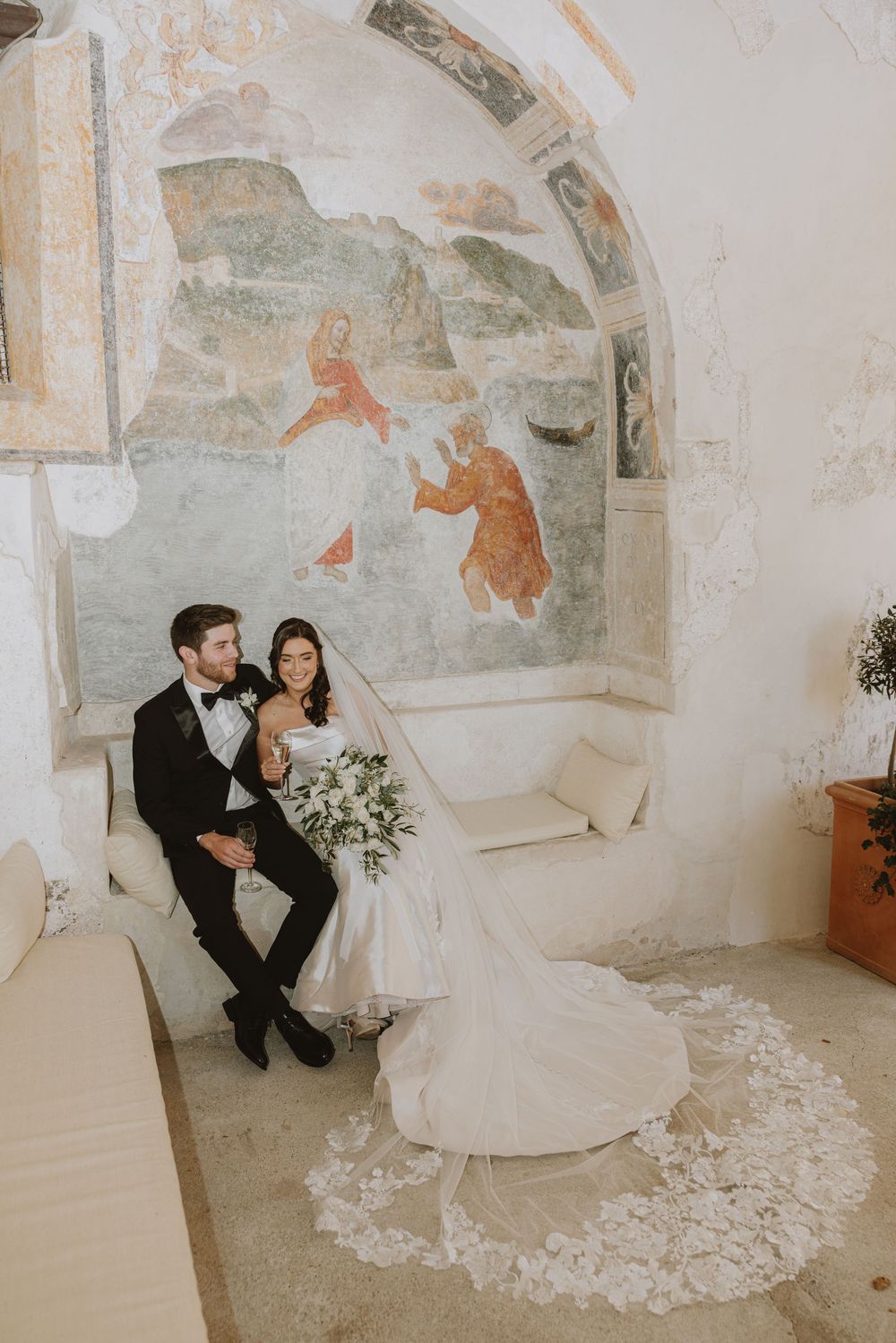 A couple sits on a white bench beneath historic frescoes in an elegant wedding setting.