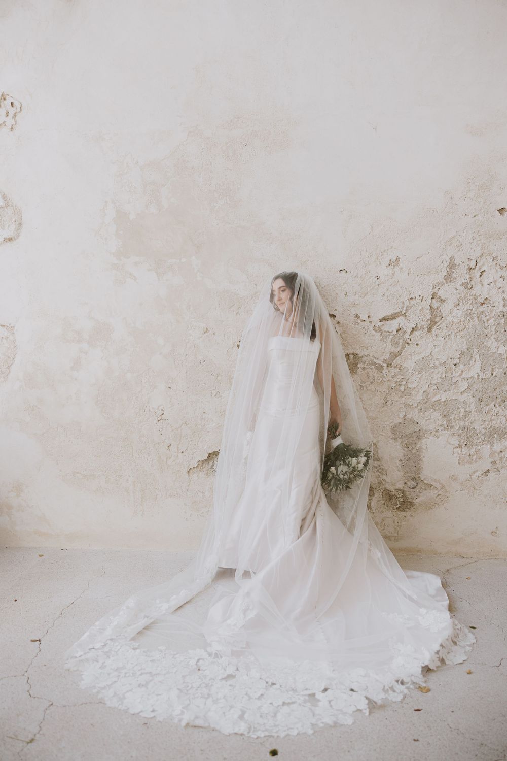A flowing white wedding dress with long veil photographed against a textured stone wall.