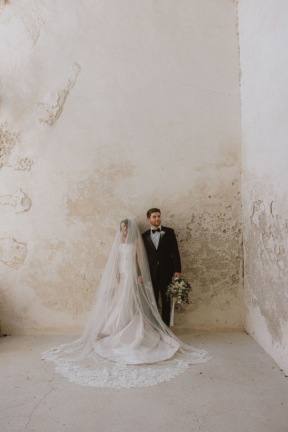 A wedding couple poses against a weathered stone wall showcasing the bride's flowing veil and train.