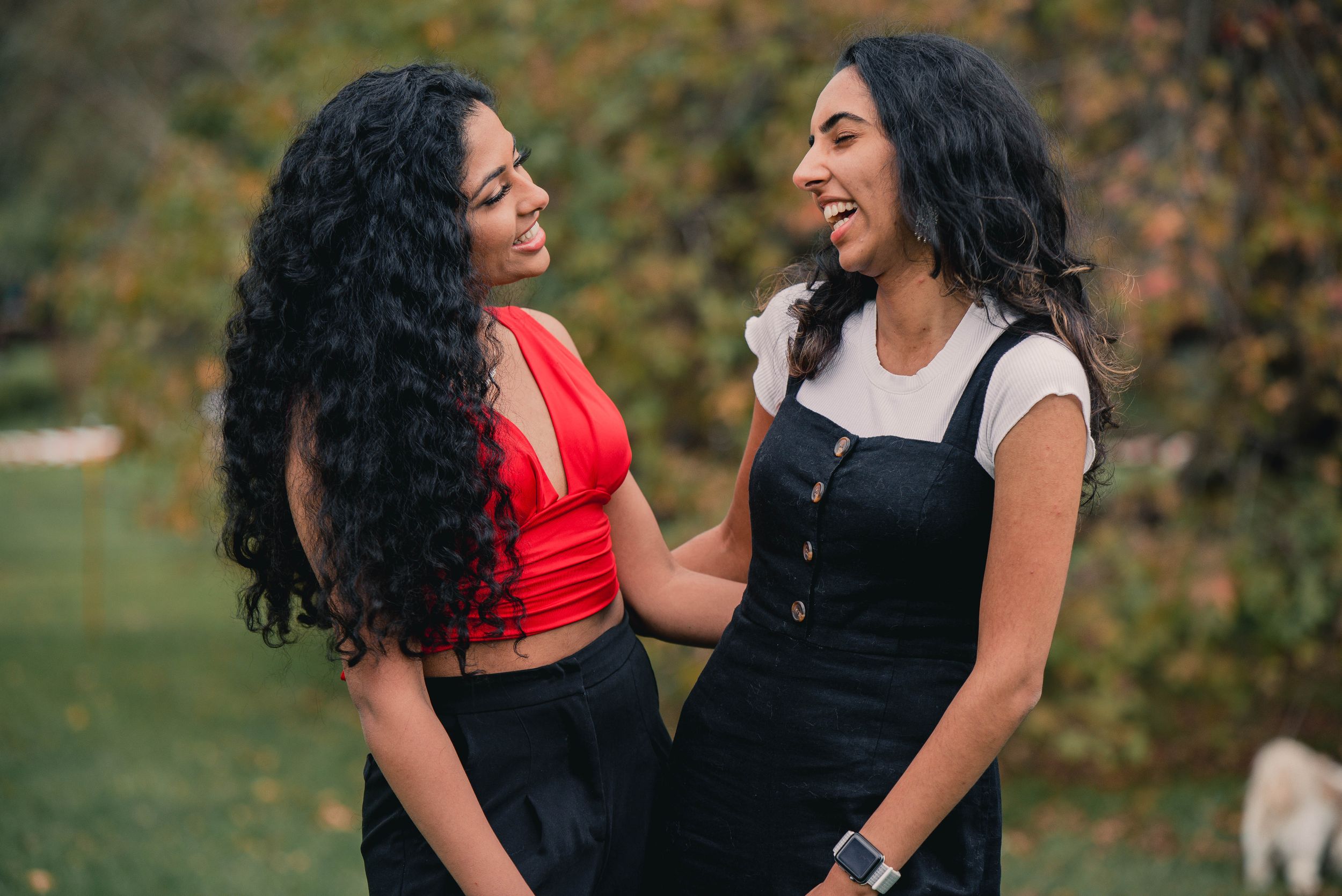 Two people with long dark curly hair share a joyful moment outdoors in casual attire against autumn foliage.