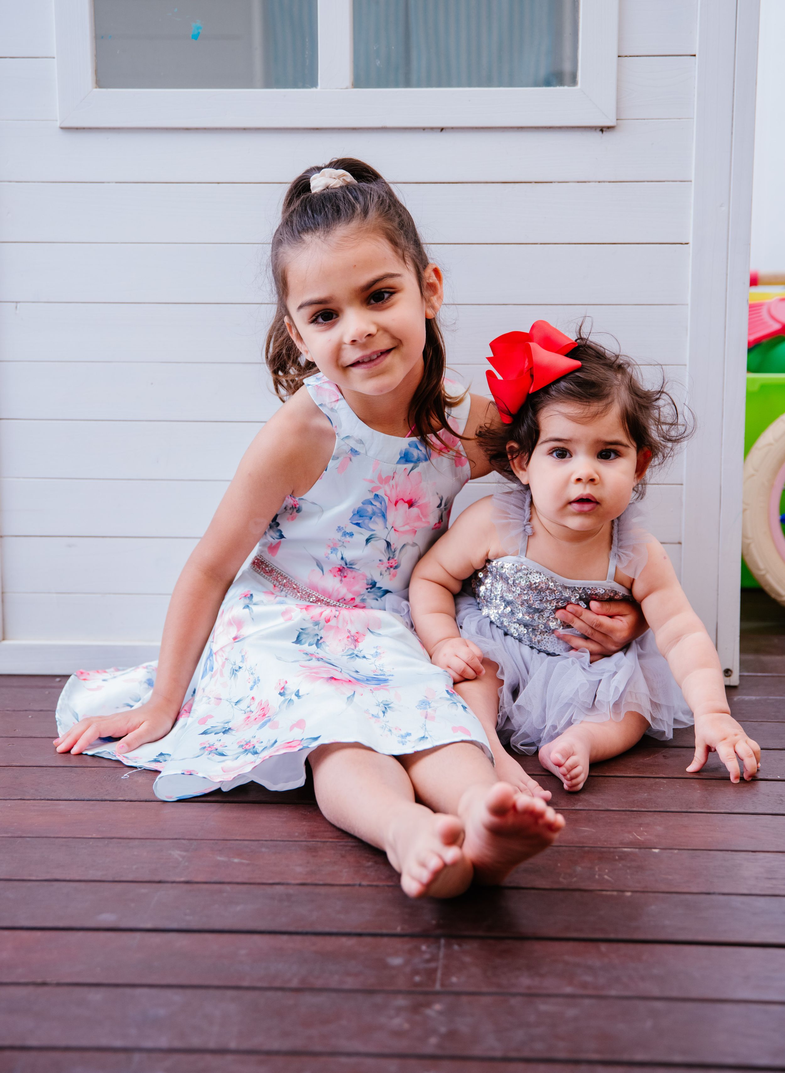 Children sitting on a wooden deck in matching floral dresses create a sweet candid moment during a summer photo session.