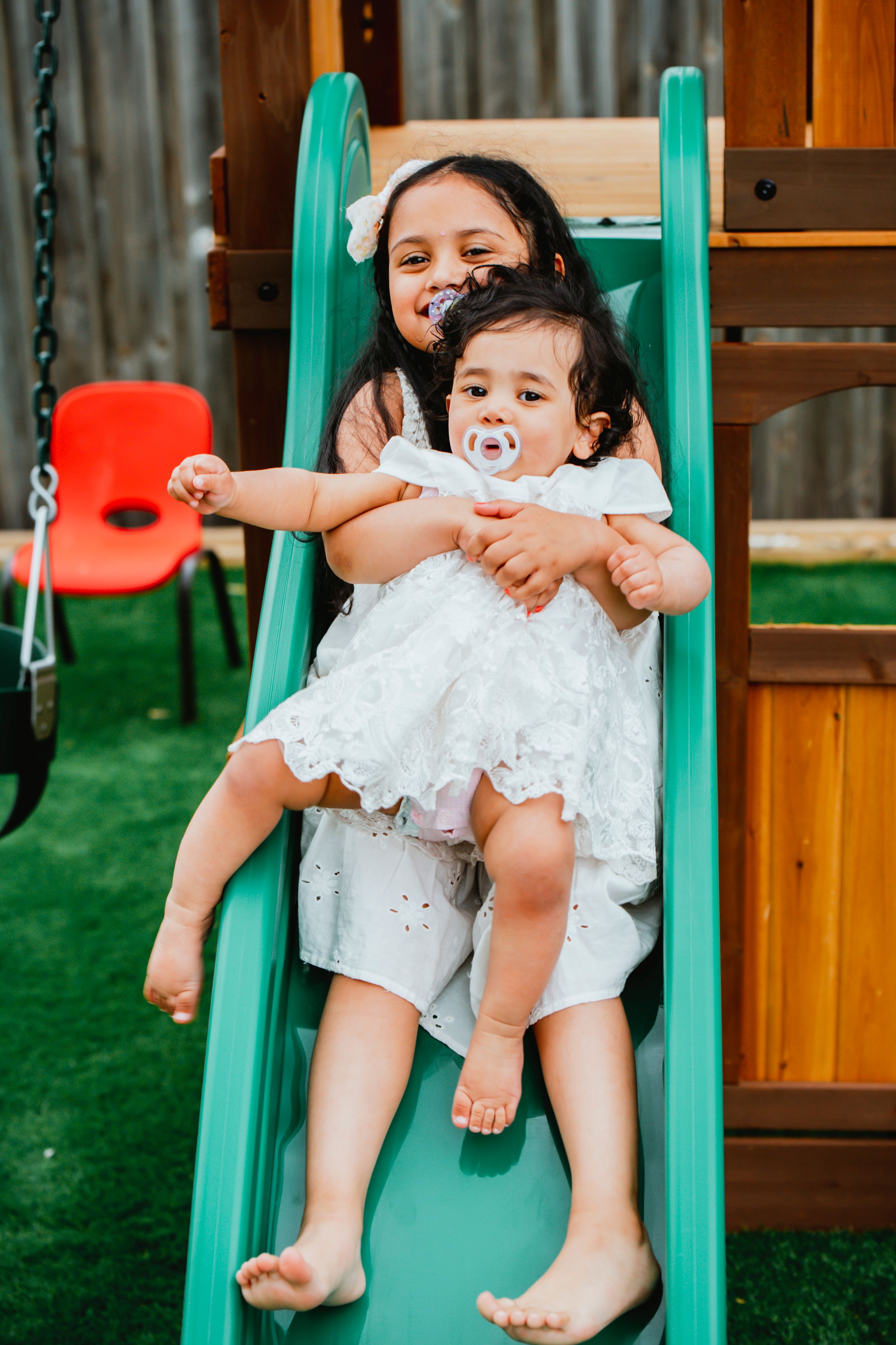 Baby in white lace dress sits on green playground swing with bare feet dangling.