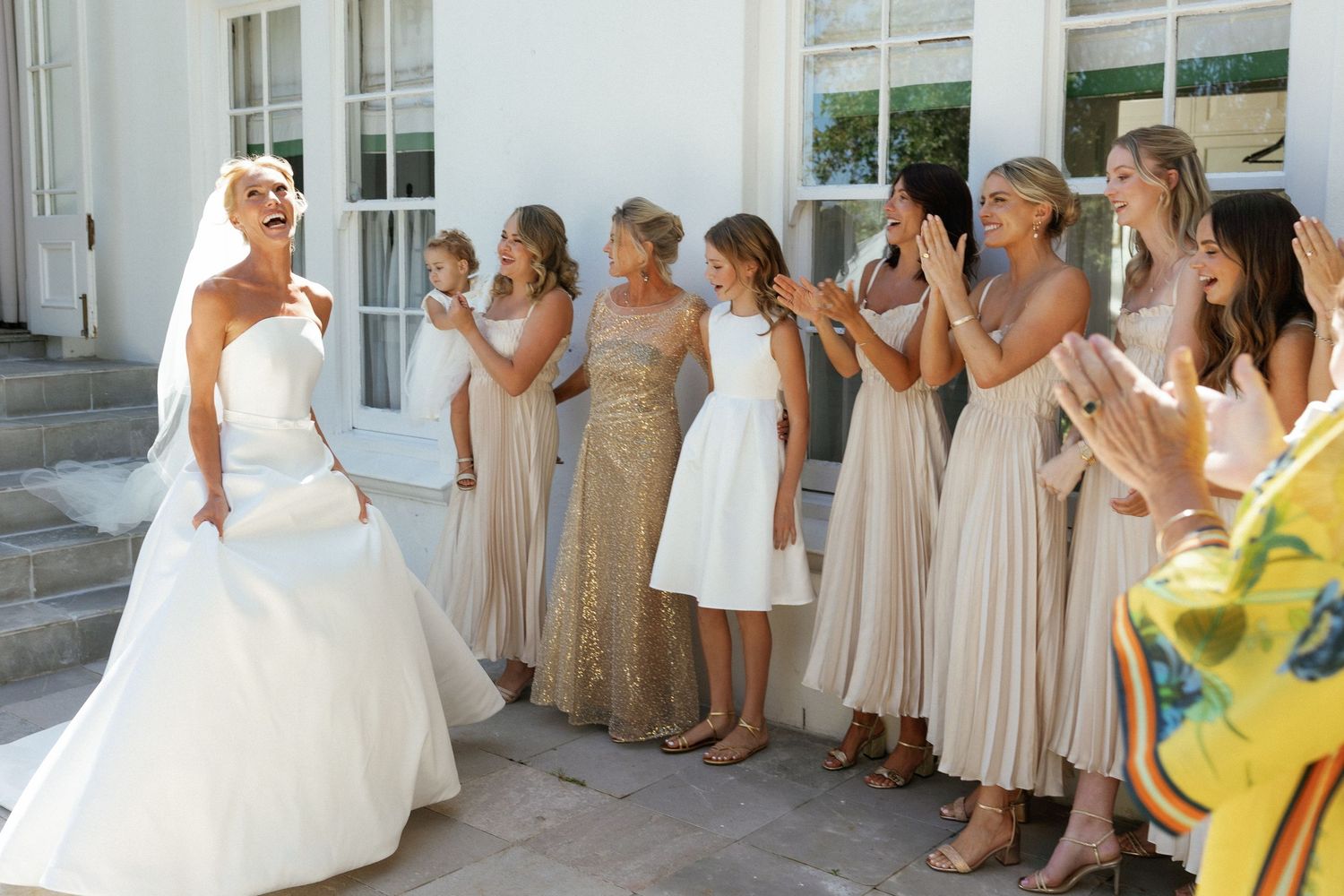 A wedding party in neutral champagne dresses poses for photos outside a white building.