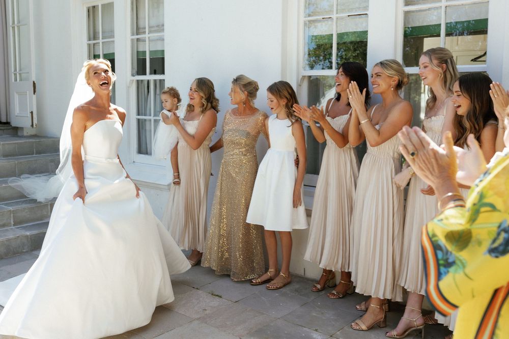 A wedding party in neutral champagne dresses poses for photos outside a white building.