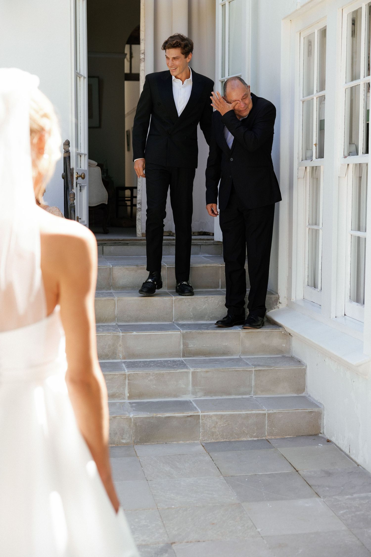 Men in formal black suits peek around a corner at someone in a white dress on stone steps.
