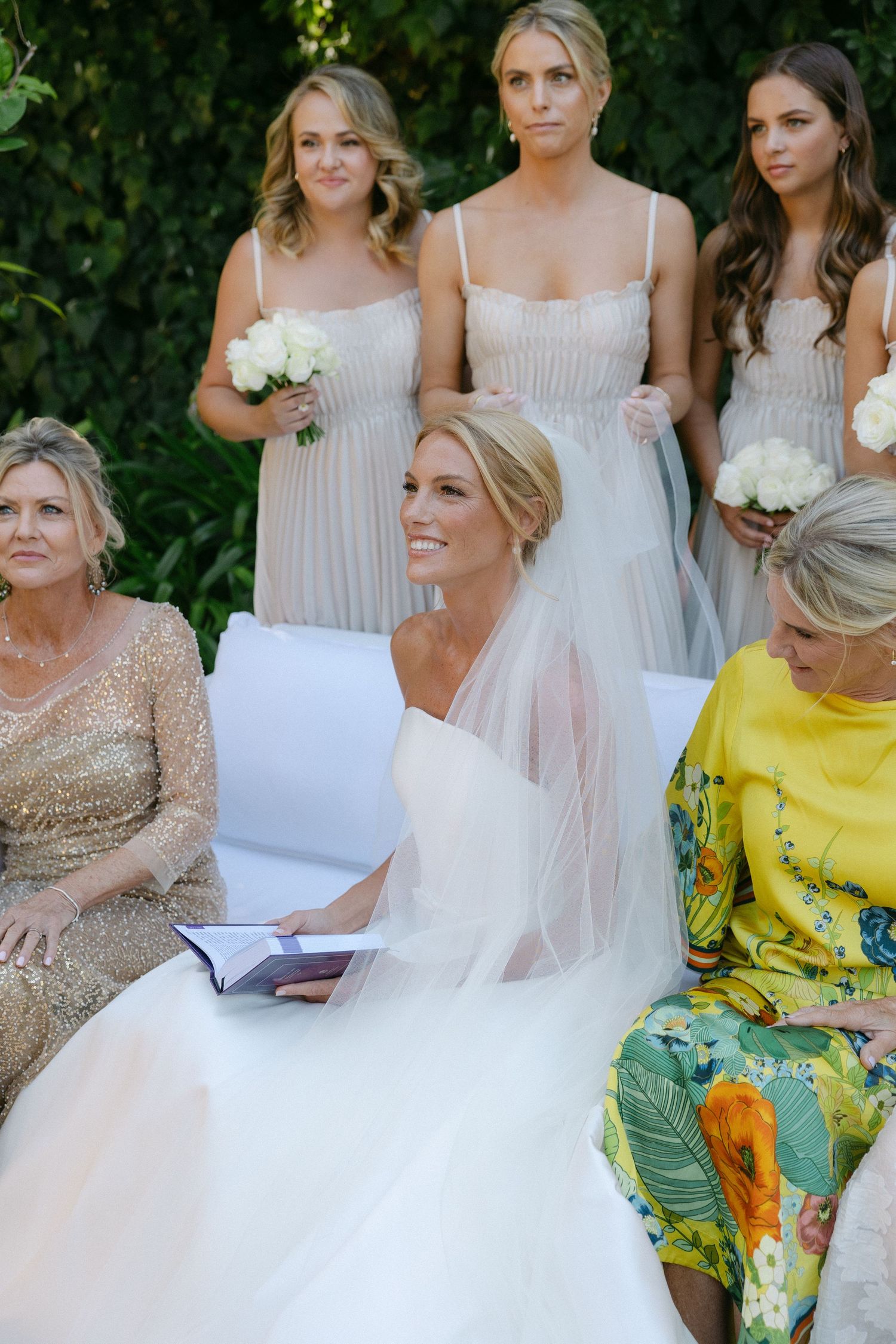 Wedding party sits outdoors with bridesmaids in light dresses and guests in colorful floral attire.