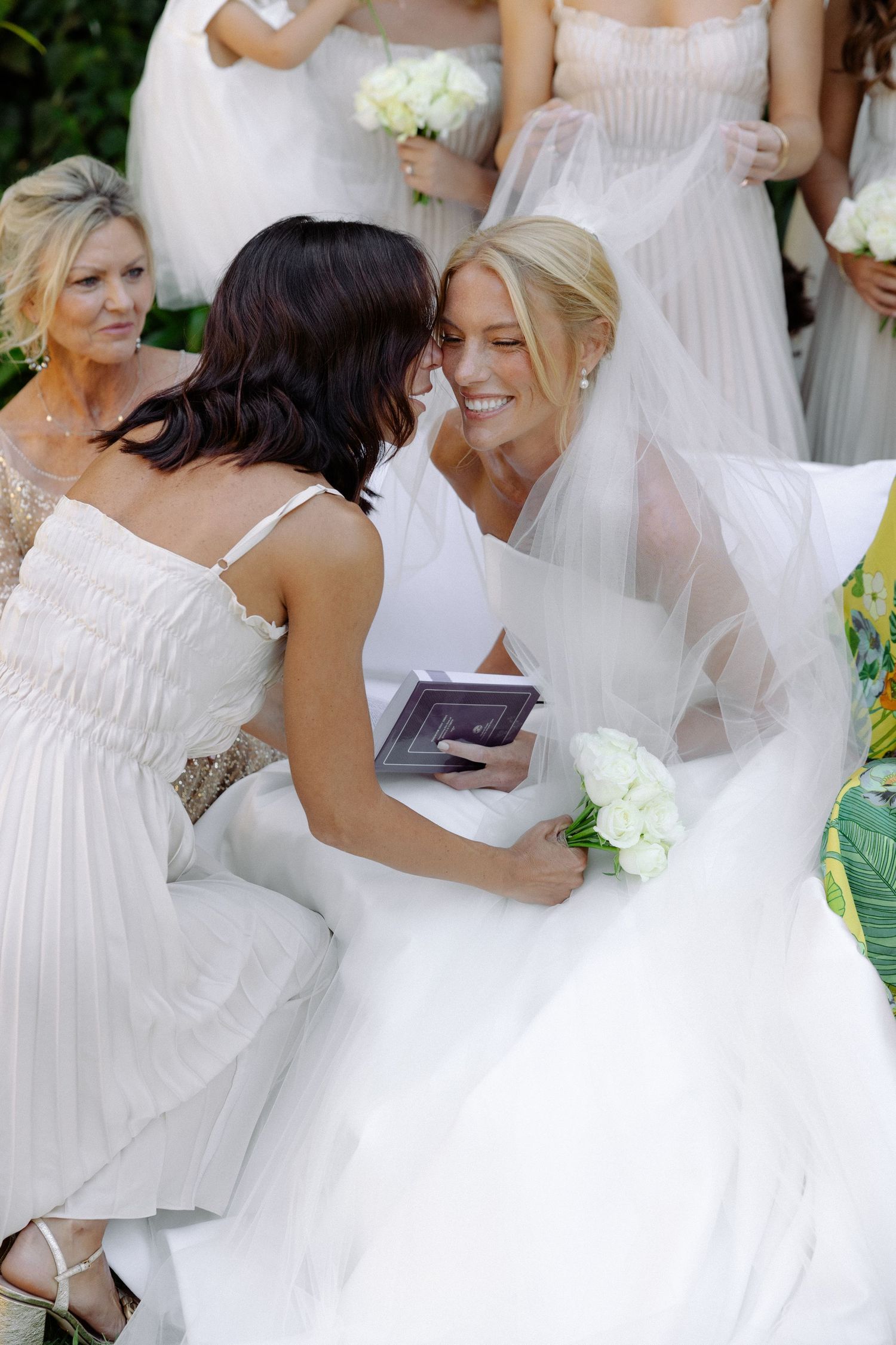Bridal party gathers outdoors in white dresses holding bouquets during a summer wedding celebration.