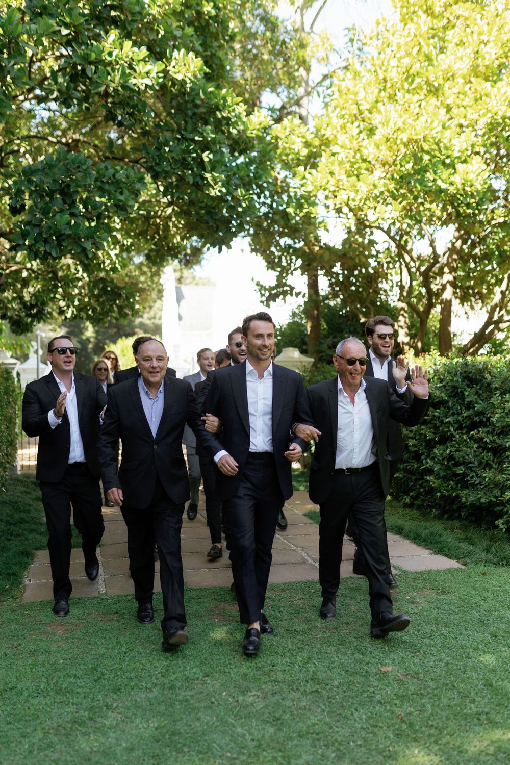 A group of well-dressed men walking together on a garden path surrounded by lush greenery.