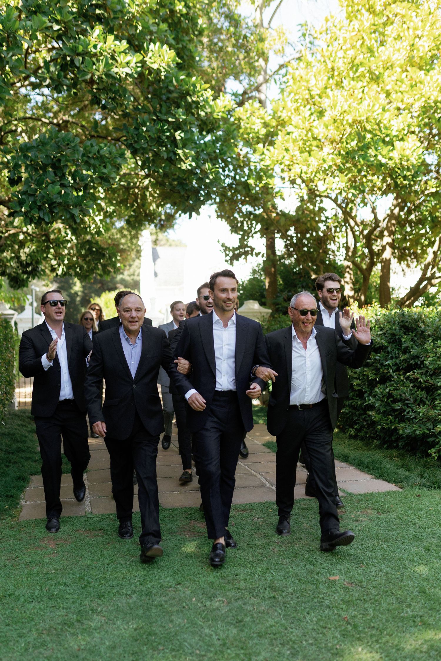 A group of well-dressed men walking together on a garden path surrounded by lush greenery.